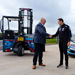 Two men shaking hands in front of a moffett forklift and Ford sports car
